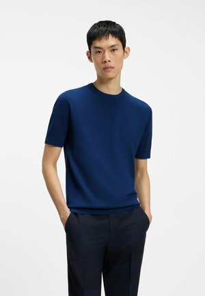 Homme aux cheveux courts et noirs portant une chemise bleu marine à manches courtes et un pantalon sombre, debout les mains dans les poches sur fond blanc.