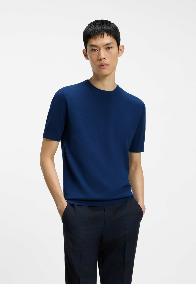 Homme aux cheveux courts et noirs portant une chemise bleu marine à manches courtes et un pantalon sombre, debout les mains dans les poches sur fond blanc.