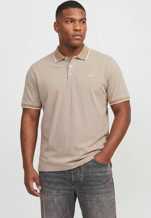 Homme en polo beige à manches courtes avec bordure blanche et jean gris, debout avec une main dans la poche, regardant légèrement sur le côté.