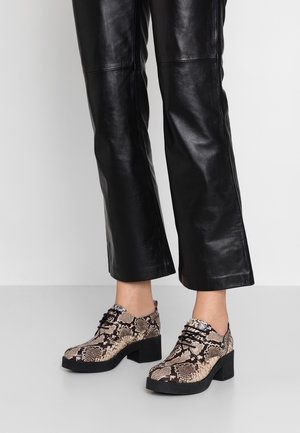 Sko med slangeskinds mønster i beige og sort, med en rund tå, flade snørebånd og en kraftig blokhæl. Parret med sorte læderbukser.