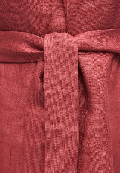 Κοντινό πλάνο δεμένης ζώνης από ύφασμα σε αχνό ρόδινο χρώμα πάνω σε αντίστοιχο υφασμάτινο ένδυμα, που αναδεικνύει τον κόμπο και τις λεπτομέρειες της ύφανσης του υφάσματος.