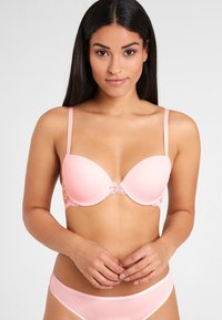 Pink padded bra with lace trim, featuring adjustable straps and a bow detail at the center. Smooth texture and rounded shape.