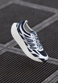 Hvide og marineblå sneakers med tyk hvid sål, detaljeret design og snørebånd, svævende over en metalgitteroverflade.