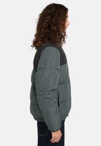Persona con capelli ricci che indossa un giubbotto invernale a due toni grigio e nero, rivolto di lato su uno sfondo bianco uniforme.