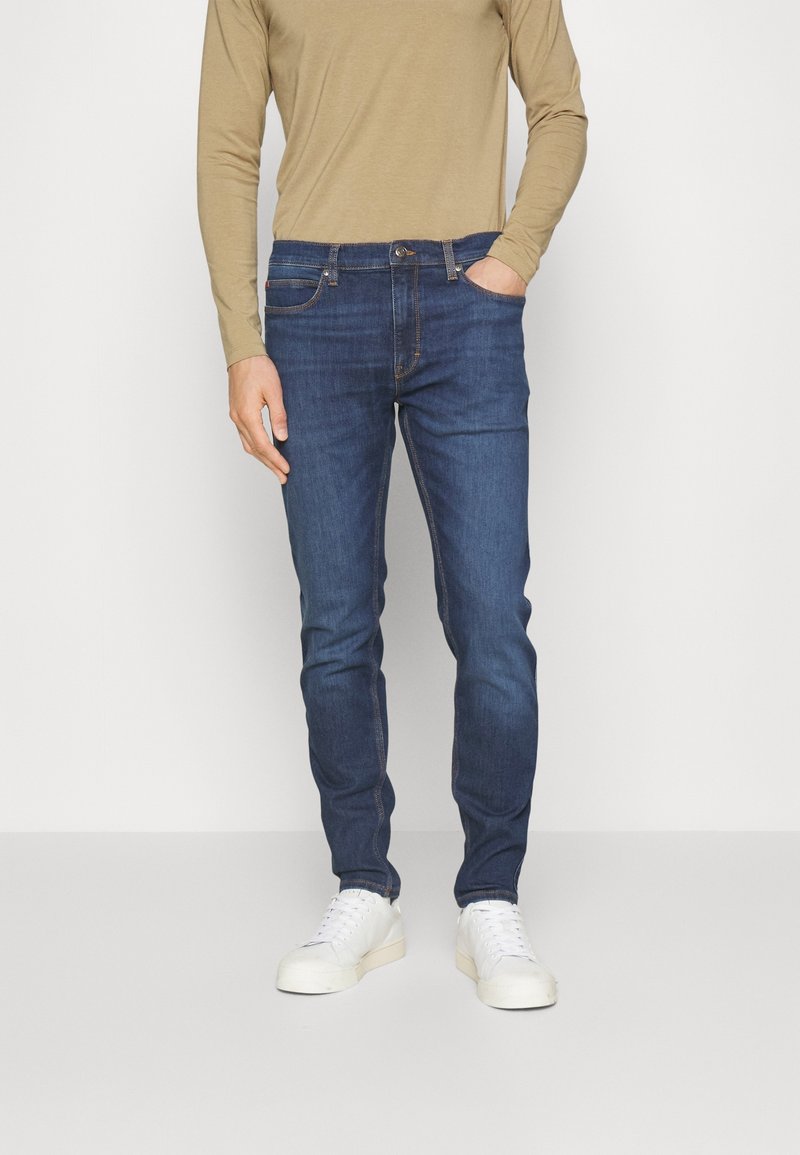 Homme portant un jean slim bleu, une chemise beige à manches longues et des baskets blanches, debout avec une main dans la poche, devant un fond uni.