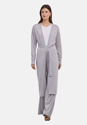 Vrouw draagt een lichtgrijze, met riem gesloten lange cardigan over bijpassende, wijde broekspijpen en een witte top, staand tegen een witte achtergrond.