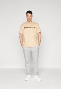 Beige Baumwoll-T-Shirt mit schwarzem "Champion"-Logo, kombiniert mit grauen Jogginghosen und weißen Sneakers. Das Model steht mit den Händen in den Taschen.