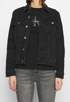 Person iført en sort denimjakke over en sort Calvin Klein T-shirt og grå jeans, med jakke-knapper delvist knappet.