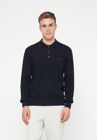 Granatowy sweter dziany z wzorem w warkocze, z kołnierzykiem i trzema guzikami. Posiada mały akcent z logo blisko klatki piersiowej. Nosi się go z jasnymi spodniami.