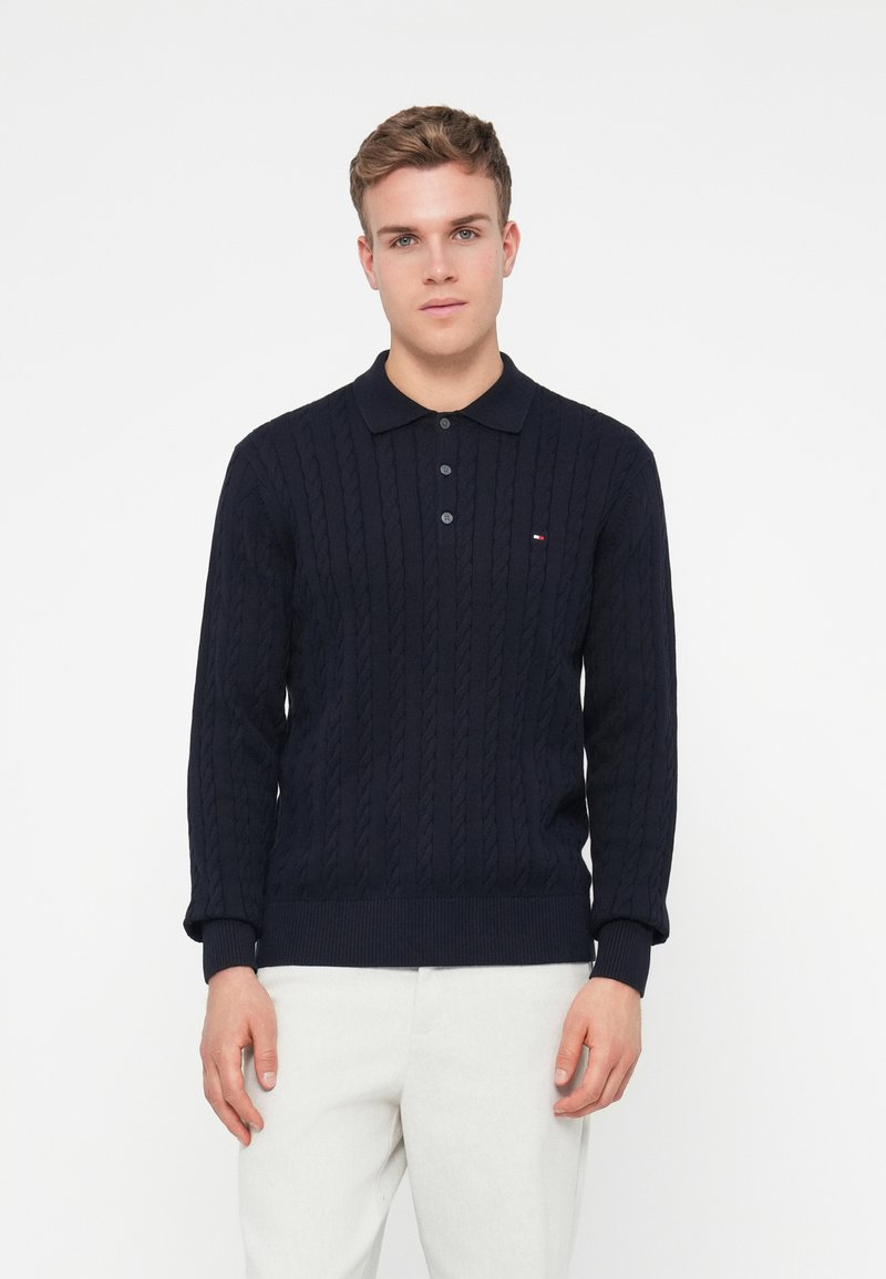 Granatowy sweter dziany z wzorem w warkocze, z kołnierzykiem i trzema guzikami. Posiada mały akcent z logo blisko klatki piersiowej. Nosi się go z jasnymi spodniami.