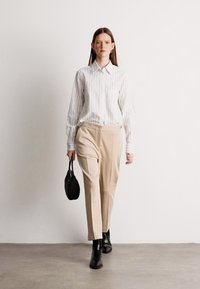 Camisa branca e riscada com botões, calças beijas com padrão, botas pretas até ao tornozelo e uma pequena mala preta. Modelo a andar contra um fundo neutro.