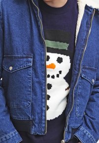 Chaqueta de mezclilla con cierre de cremallera, de color azul, con dos bolsillos en el pecho. Debajo, un suéter azul marino presenta un diseño de muñeco de nieve con nariz de zanahoria naranja y un sombrero verde.