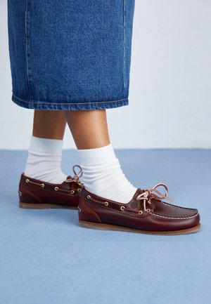 Scarpe da barca in pelle marrone con lacci bianchi, occhielli in metallo e suola texturizzata, abbinate a calzini bianchi e una gonna di denim blu.