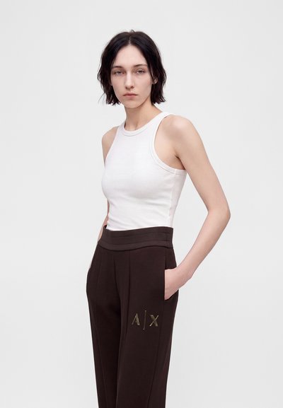 Jeune femme aux cheveux courts et foncés porte un haut blanc sans manches et un pantalon foncé avec le logo A|X, mains dans les poches, expression neutre, fond uni.