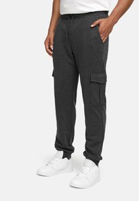 Pantalon de jogging en coton gris avec poches latérales et poches cargo sur les jambes, doté d'une taille avec cordon de serrage et de poignets élastiques. Porté avec des baskets blanches.