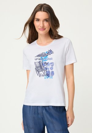 Frau trägt ein weißes T-Shirt mit Strandstuhl, Sonnenschirm, Sonnenbrille, Blumen und dem Schriftzug „Beach Vibes“ kombiniert mit blauer Hose.