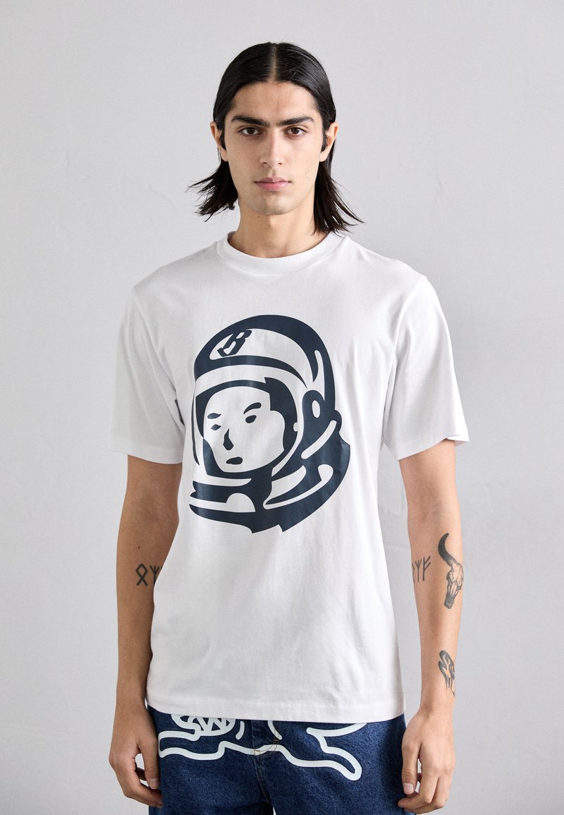 White cotton t-shirt featuring a large navy astronaut graphic on the front. Model is wearing blue jeans with white accents.