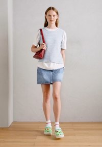 Blaues, ärmelloses Stricktop über einem weißen Hemd, kombiniert mit einem Jeans-Minirock und grünen Clogs mit Cartoon-Motiven sowie einer roten Handtasche.