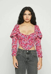 Mulher com longos cabelos pretos encaracolados, vestindo um top corselet vermelho com mangas bufantes florais e jeans pretos de cintura alta, de pé contra um fundo liso.