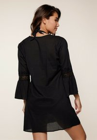 Vestido negro con mangas acampanadas de tres cuartos, que presenta detalles de encaje en las mangas y una espalda descubierta con un lazo de cierre. Material ligero.