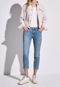 Giacca di jeans beige chiaro con bottoni a pressione, indossata sopra una camicia bianca. Jeans blu sbiaditi, tagliati alla caviglia, abbinati a sneaker bianche.