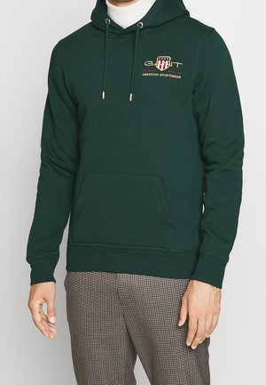 Man wearing dark green hooded sweatshirt with front pocket and "GANT American Sportswear" logo, paired with checkered pants.