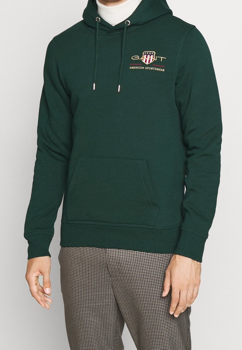 Homme portant un sweat à capuche vert foncé avec poche avant et logo « GANT American Sportswear », associé à un pantalon à carreaux.