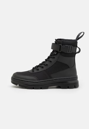 Botte montante noire avec fermeture à lacets à l’avant, semelle épaisse, lanière à la cheville avec boucle et languette à l’arrière sur un fond clair uni.