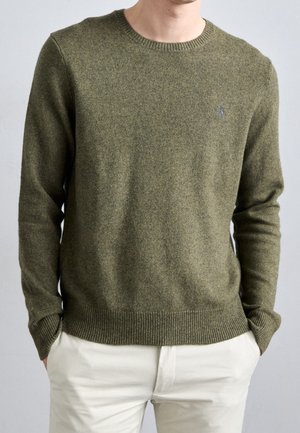 Mann trägt einen olivgrünen gestrickten Pullover mit gerippten Bündchen und Saum, kombiniert mit hellbeigen Hosen, die Hände in den Taschen.