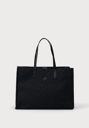 Svart tote bag med kviltet tekstur, med to solide håndtak og en liten metallisk logo foran. Rektangulær form.