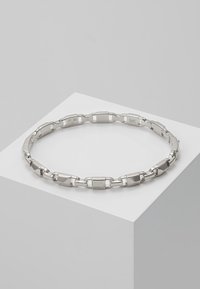 Bracelet en argent composé de segments métalliques polis rectangulaires et carrés, posé sur un piédestal géométrique blanc sur fond gris.