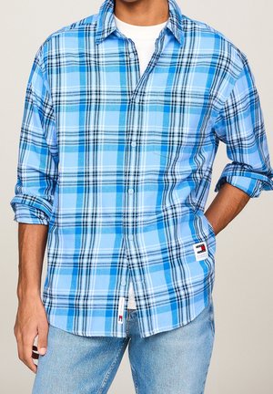 Man draagt een lichtblauw, wit en zwart geruit overhemd met knopen over een wit T-shirt met opgerolde mouwen en lichtblauwe spijkerbroek.