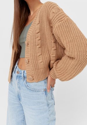 Cardigan beige en tricot avec motif de torsades, manches larges et trois boutons, associé à un jean taille haute bleu clair.