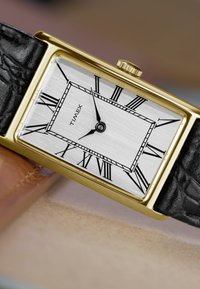 Goldene rechteckige Timex-Armbanduhr mit schwarzen römischen Ziffern, schwarzen Stunden- und Minutenzeigern und schwarzem Lederarmband auf beigem Untergrund.