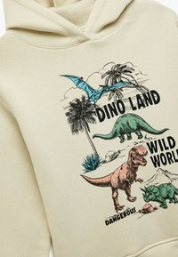 Sudadera color crema con ilustraciones de dinosaurios en verde y rosa, palmeras y el texto "DINO LAND WILD WORLD DANGEROUS". Textura suave.