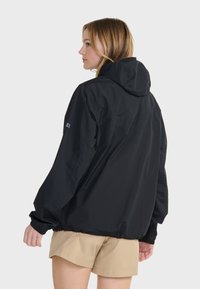 Veste noire à capuche avec une coupe ample, des poignets élastiques et une texture lisse. Comprend un détail de logo sur la manche. Portée par-dessus des shorts beiges.