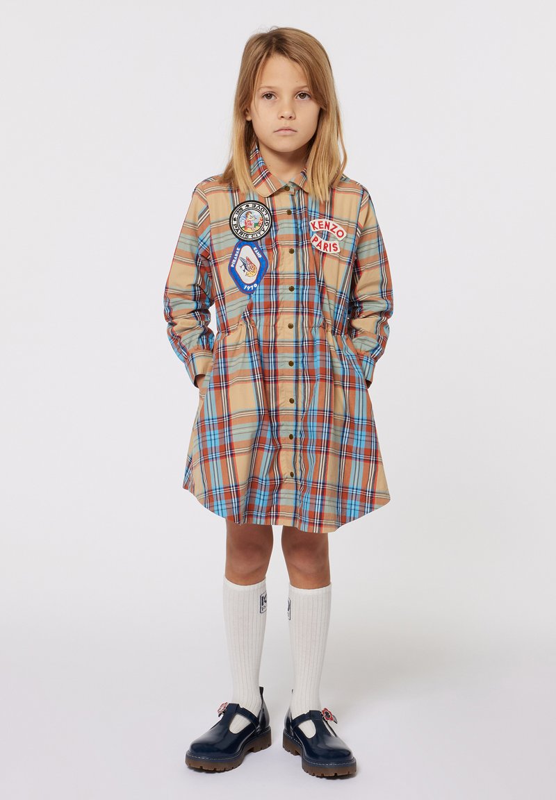 Abito a camicia a quadri nei toni beige, blu e arancione, con toppe. Abbinato a calze bianche al ginocchio e scarpe navy.