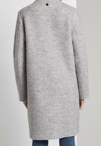 Grå ullrock med texturerad yta, långa ärmar och hög krage. Har en svart knapp på baksidan och når till mitten av låret.