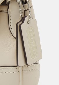 Beige leather handbag with a textured finish, featuring a dark brown accent and two attached tags. Detailed stitching along the seams.