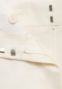 Gros plan sur la ceinture d’un pantalon blanc montrant un bouton, des crochets métalliques et des couches de tissu superposées pour la fermeture.