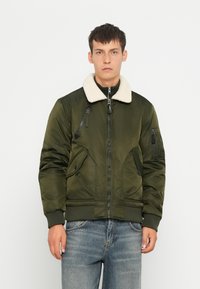 Veste bomber verte avec col en shearling, fermeture éclair, plusieurs poches zippées et poignets côtelés. Portée par-dessus une chemise à accent sombre et un jean.
