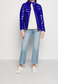 Blaue, glänzende Daunenjacke mit Reißverschluss vorne, hohem Kragen und gepolstertem Design. Getragen über einem leichten Pullover und kombiniert mit hellen, ausgestellten Jeans.