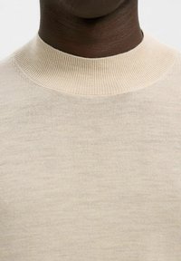Pull en beige à col montant avec un col côtelé, fabriqué en tissu lisse et léger. Présente un design épuré et minimaliste sans motifs.