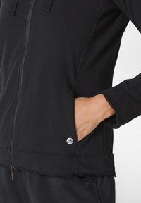 Giacca nera con zip realizzata in tessuto morbido, dotata di tasca a marsupio e dettagli con logo. Texture liscia, vestibilità atletica e maniche lunghe.