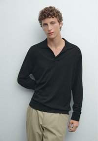 Svart långärmad polo med V-ringad krage och avslappnad passform, ihop med beigea byxor, mot en neutral bakgrund.