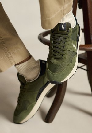 Noge u maslinasto zelenim tenisicama s vezicama, bijelim potplatima i Polo logom, krem hlače, stoje na prečki drvenog stolca.