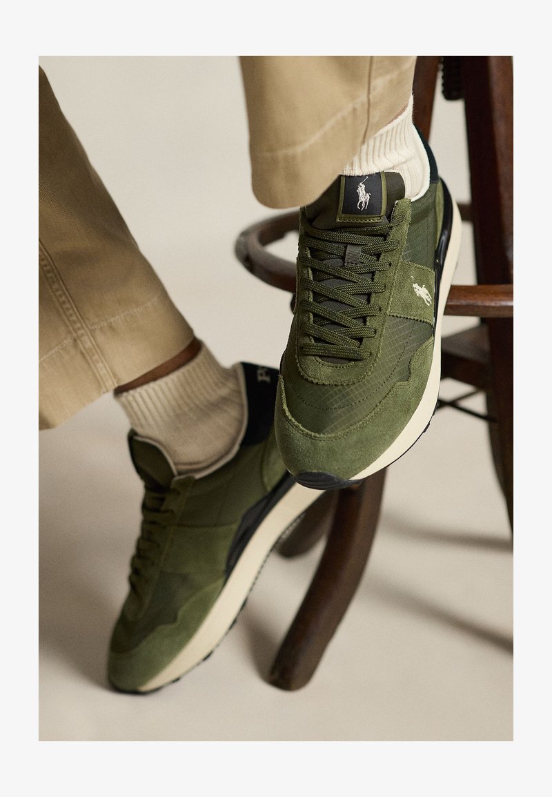 Kojos su alyvų žalių raišteliais batelių su baltais padais ir Polo logotipu, smėlio spalvos kelnės, stovinčios ant medinės taburetės laiptelio.