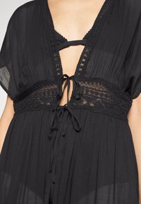 Prenda negra con un profundo escote en V, detalles de crochet en la cintura y lazos para ajuste. Tejido suave y ligero con una textura fluida.