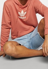 Hoodie i korall med vit Adidas-logga; bärs med ljusblå slitna denimshorts. Mjuk textur, avslappnad passform.