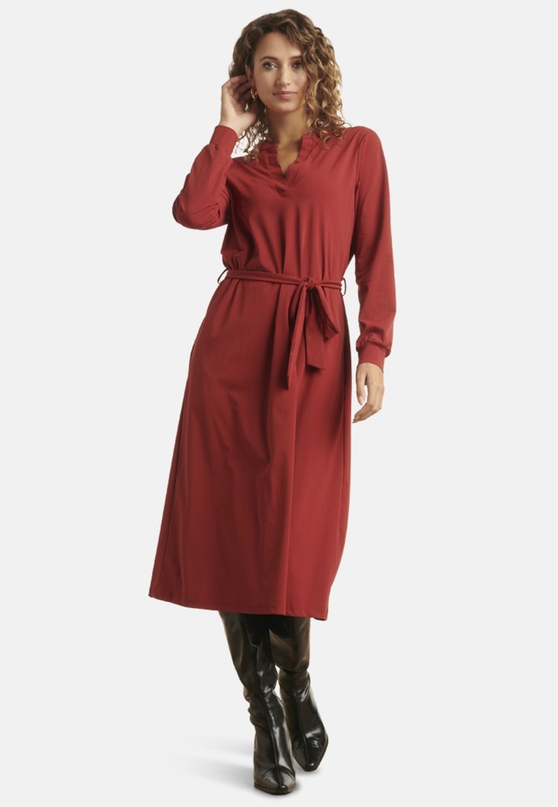 Red long-sleeve dress with a v-neck and cinched waistline, featuring a tie belt and flowing fabric, paired with black knee-high boots.